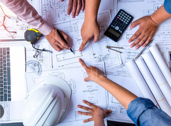 Plusieurs mains autour de plans techniques, avec un casque de chantier, une calculatrice et des rouleaux de plans